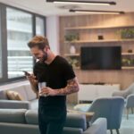 a man in a black shirt standing in his living room playing Pokemon TCG Pocket on his phone image source: https://www.pexels.com/photo/man-in-black-shirt-standing-while-holding-drinking-glass-942424/