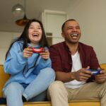 a couple sitting on a couch in their home holding Playstation 5 controllers and playing Split Fiction